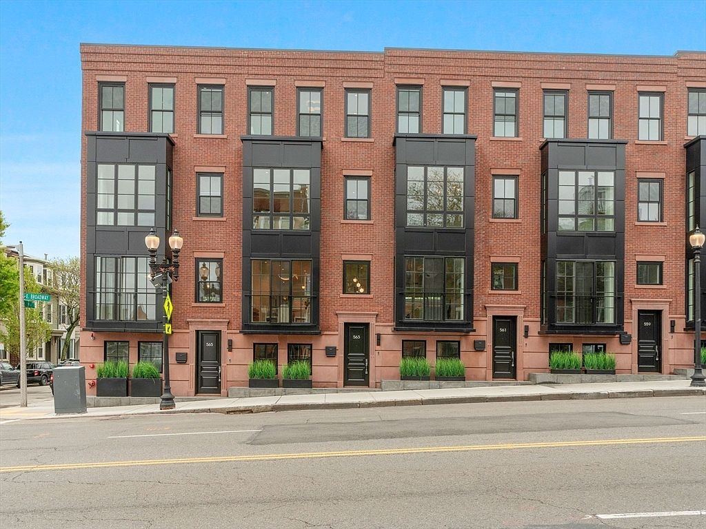 The image showcases the front facade of a modern brick townhouse complex. The buildings feature a blend of traditional brickwork with contemporary black window frames and protruding bay windows. The street is visible in the foreground, adding context to the urban setting.
