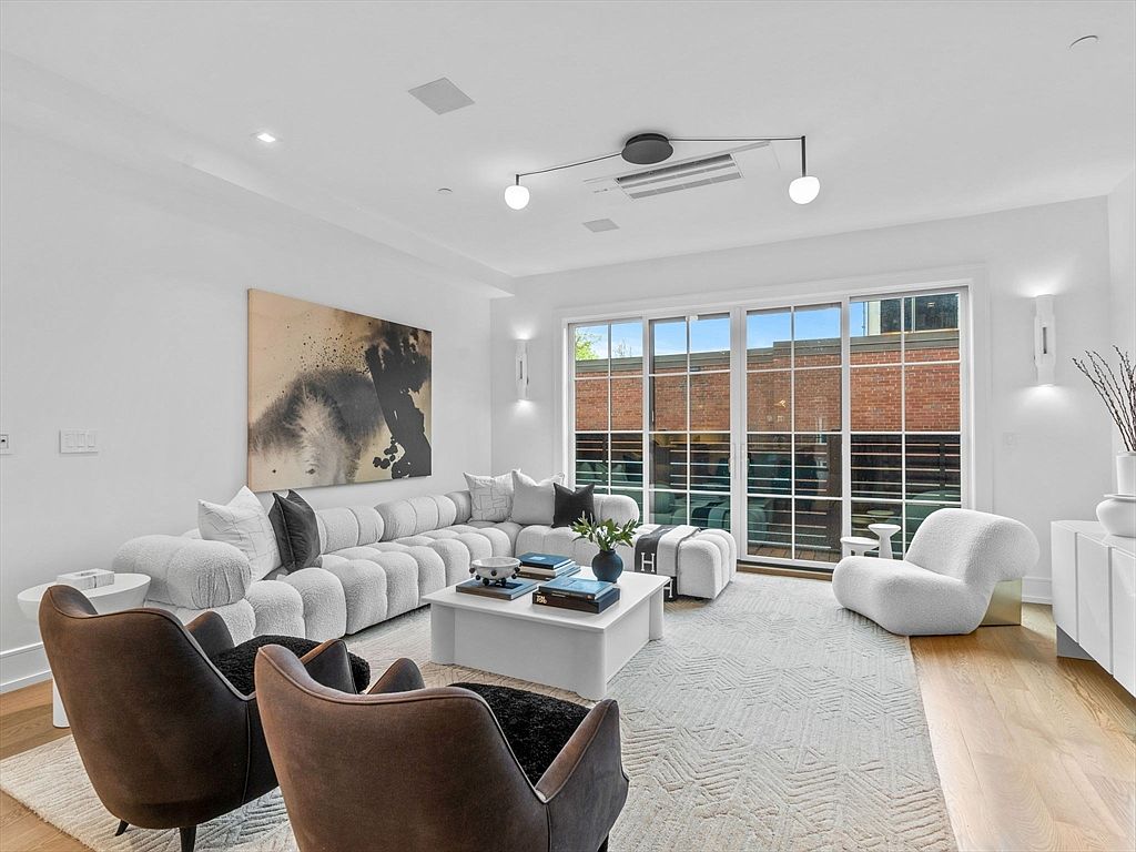 This is a bright and modern living room featuring a large, modular white sofa, two brown armchairs, and a white coffee table. A large abstract painting hangs on the wall, and a wall of windows provides natural light and a view of the exterior. The room is decorated in a minimalist style with a neutral color palette, creating a sophisticated and inviting atmosphere.
