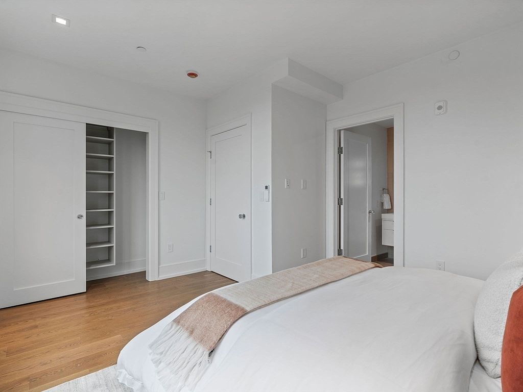 This is a bright and airy primary bedroom featuring hardwood floors and white walls. A large bed with white linens and a textured throw is the focal point, while a closet with sliding doors and built-in shelving provides ample storage. An ensuite bathroom is visible through a doorway, adding to the convenience and appeal of the space.