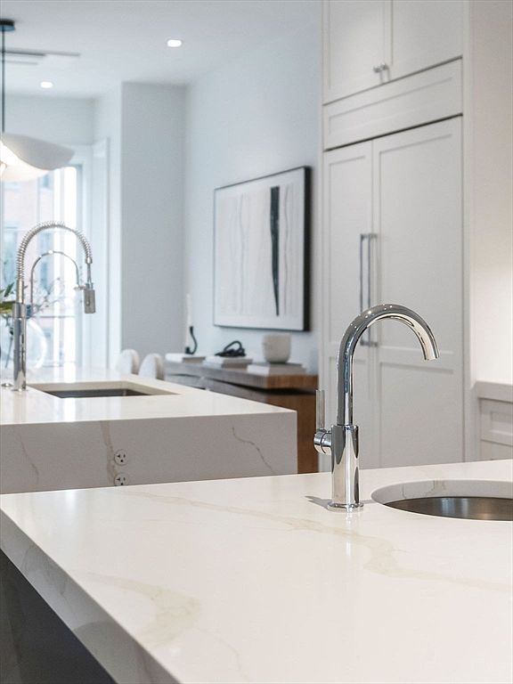 This is a bright, modern kitchen featuring white countertops with subtle veining and stainless steel fixtures. The focus is on the clean lines and minimalist design, with a partial view of a second island and built-in refrigerator. The perspective is a close-up shot, highlighting the quality of the materials and the sleek aesthetic.