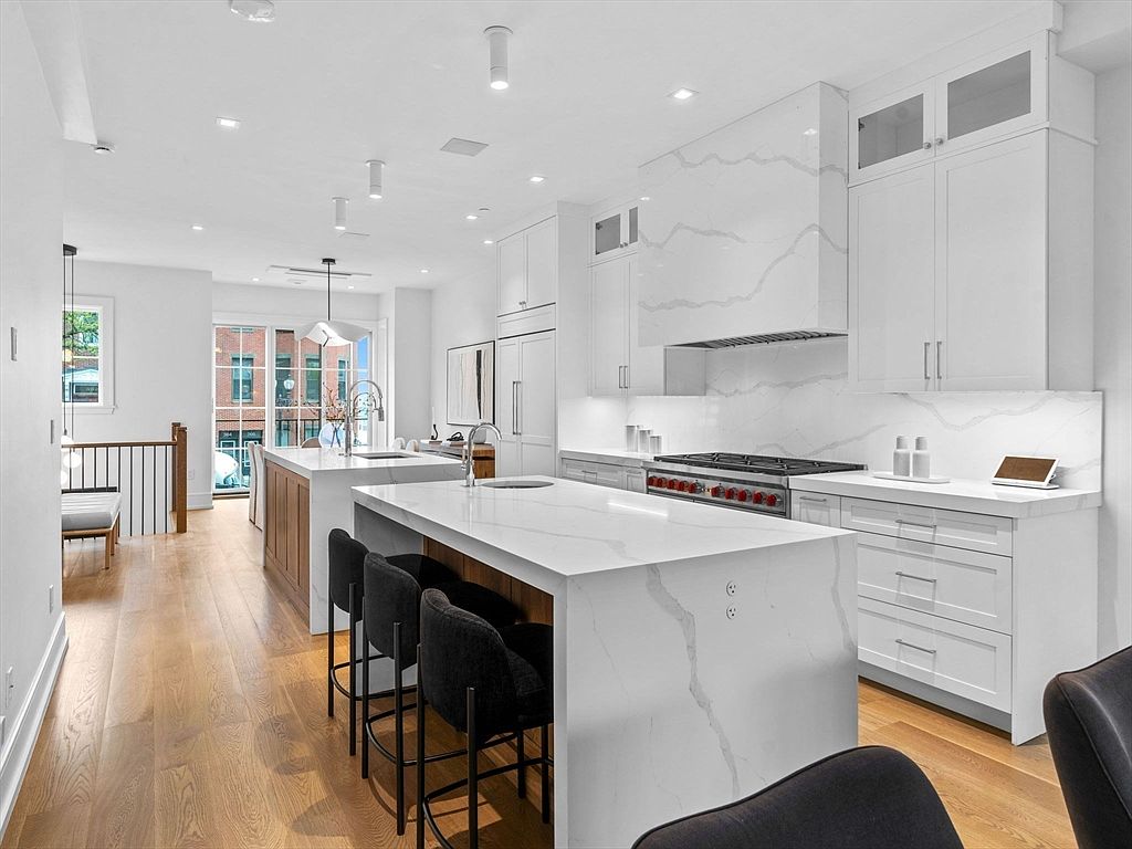 This is a bright and modern kitchen featuring a large island with seating, white cabinetry, and marble countertops. The kitchen is well-lit with recessed lighting and natural light, creating a clean and inviting atmosphere. The open layout connects seamlessly to the adjacent living space, enhancing the sense of spaciousness.