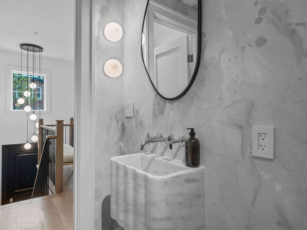 This image showcases a stylish guest bathroom featuring a marble pedestal sink with a modern chrome faucet and a dark amber soap dispenser. The wall is adorned with a unique gray marble-like texture, complemented by round sconces and an oval mirror with a black frame. The view extends to a hallway with stairs, adding depth and interest to the composition.
