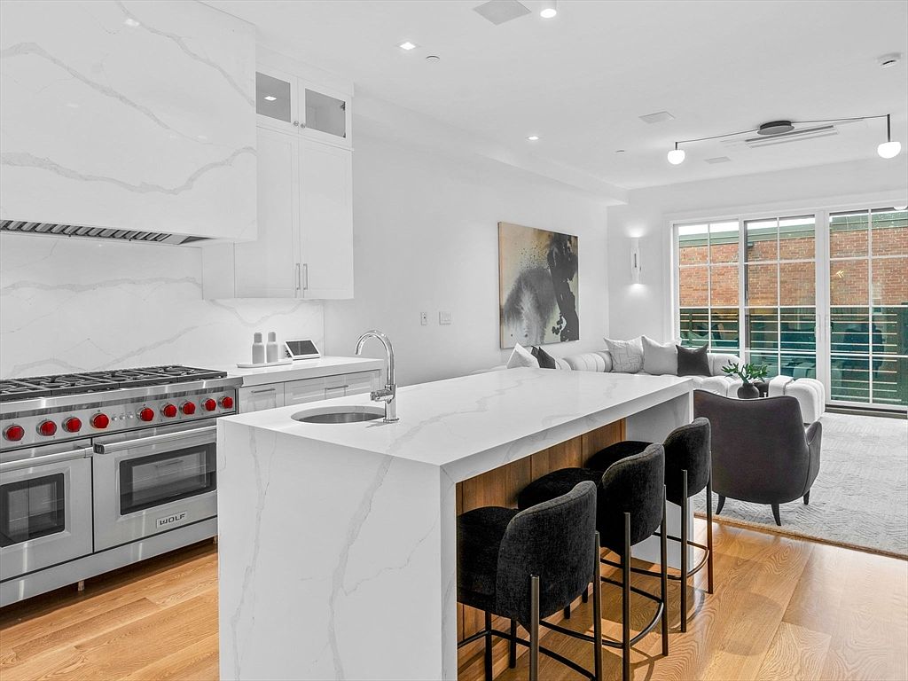 This is a bright, modern kitchen featuring a large island with a waterfall countertop and seating for three. Stainless steel appliances, including a Wolf range, are prominent, and the open floor plan seamlessly connects the kitchen to the living area. The hardwood floors and contemporary lighting fixtures add to the upscale feel of the space.