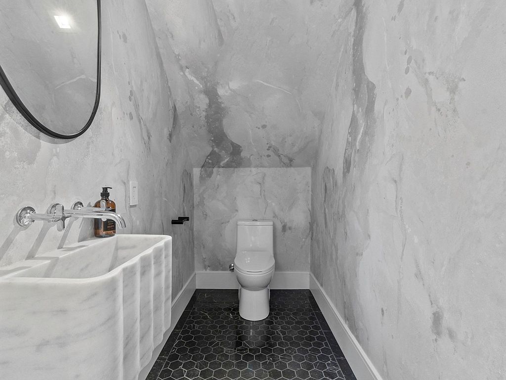 This is a modern guest bathroom featuring a unique marble sink with a wall-mounted faucet and a round mirror. The walls are covered in a marble-like wallpaper, and the floor is tiled with a dark, geometric pattern. The toilet is neatly tucked into a small alcove, creating a compact yet stylish space.