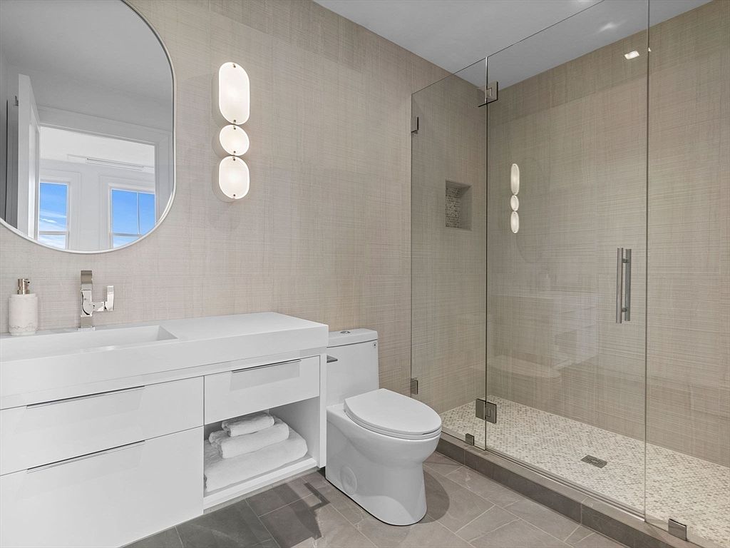 This is a well-lit primary bathroom featuring a modern aesthetic. The bathroom includes a white vanity with a sleek countertop and storage, a toilet, and a glass-enclosed shower with tiled walls and floor. The walls are covered in a textured wallpaper, and unique vertical light fixtures add a touch of elegance.