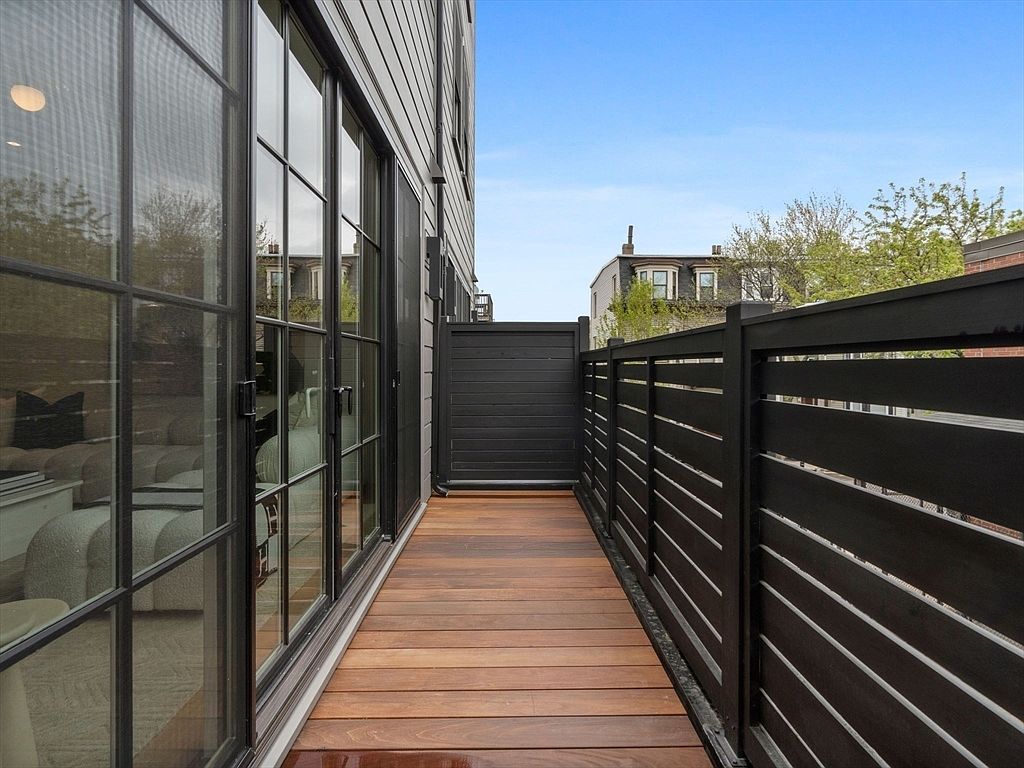 This image showcases a modern balcony or deck area featuring a sleek, dark-stained wooden floor and a contemporary black fence with horizontal slats. Large glass doors provide access to the interior, reflecting the light and suggesting a seamless indoor-outdoor transition. The overall impression is one of stylish urban living with a focus on privacy and design.