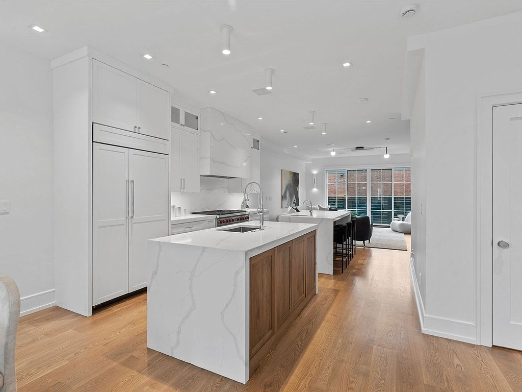 This is a bright and modern kitchen featuring a large island with a white countertop and wooden base. The kitchen is equipped with stainless steel appliances and white cabinetry, creating a clean and spacious feel. The open floor plan extends into a living area with large windows, providing ample natural light.