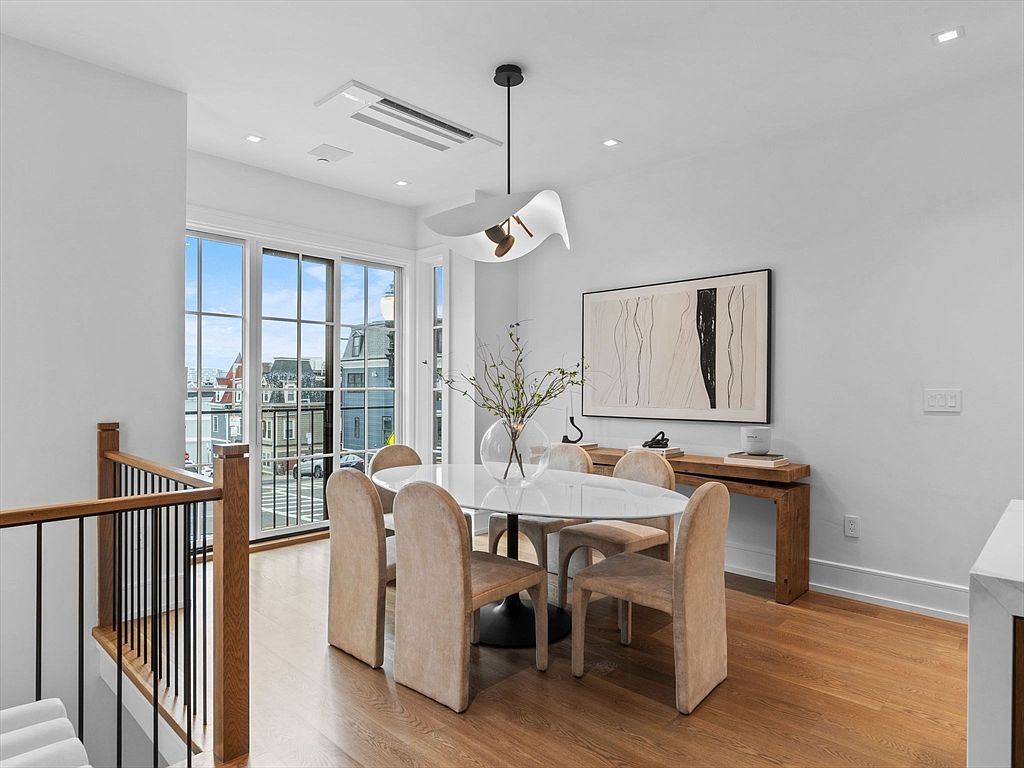 This is an interior shot of a dining room featuring a round white marble table with a dark base, surrounded by six upholstered chairs in a neutral tone. A modern chandelier hangs above the table, and a large piece of abstract art adorns the wall. The room has hardwood floors and large windows that offer a view of the neighborhood, creating a bright and inviting space.