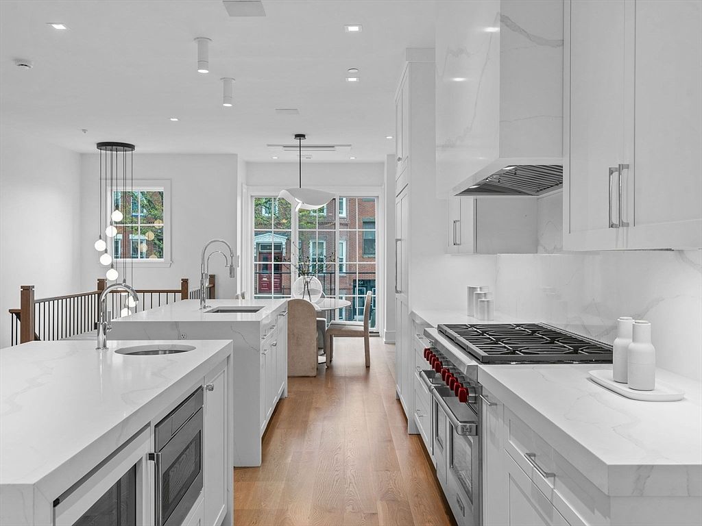 This is a bright and modern kitchen featuring white cabinetry, marble countertops, and stainless steel appliances. The kitchen includes two islands, a high-end range, and a dining area visible through a large window. The overall impression is clean, luxurious, and highly functional, perfect for a discerning buyer.