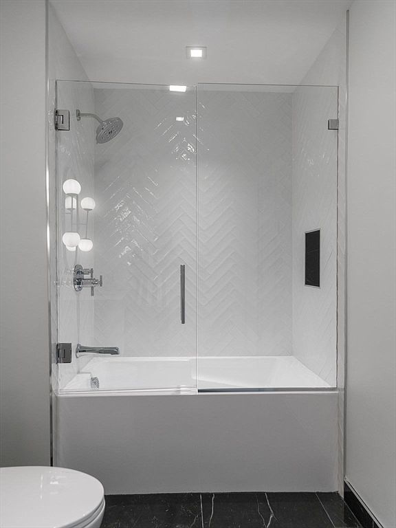 This is a well-lit primary bathroom featuring a modern bathtub and shower combination enclosed by glass doors. The walls are adorned with white herringbone tiles, and the fixtures are chrome. The overall impression is clean, bright, and contemporary, suggesting a luxurious and relaxing space.
