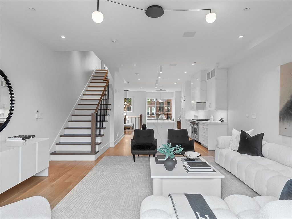 This is an interior shot of a modern living room, showcasing an open-concept design that flows into the kitchen. The room features a white sofa, black armchairs, a light gray rug, and a white coffee table with decorative items. A staircase is visible on the left, and the overall aesthetic is bright, clean, and contemporary, creating an inviting and spacious atmosphere.