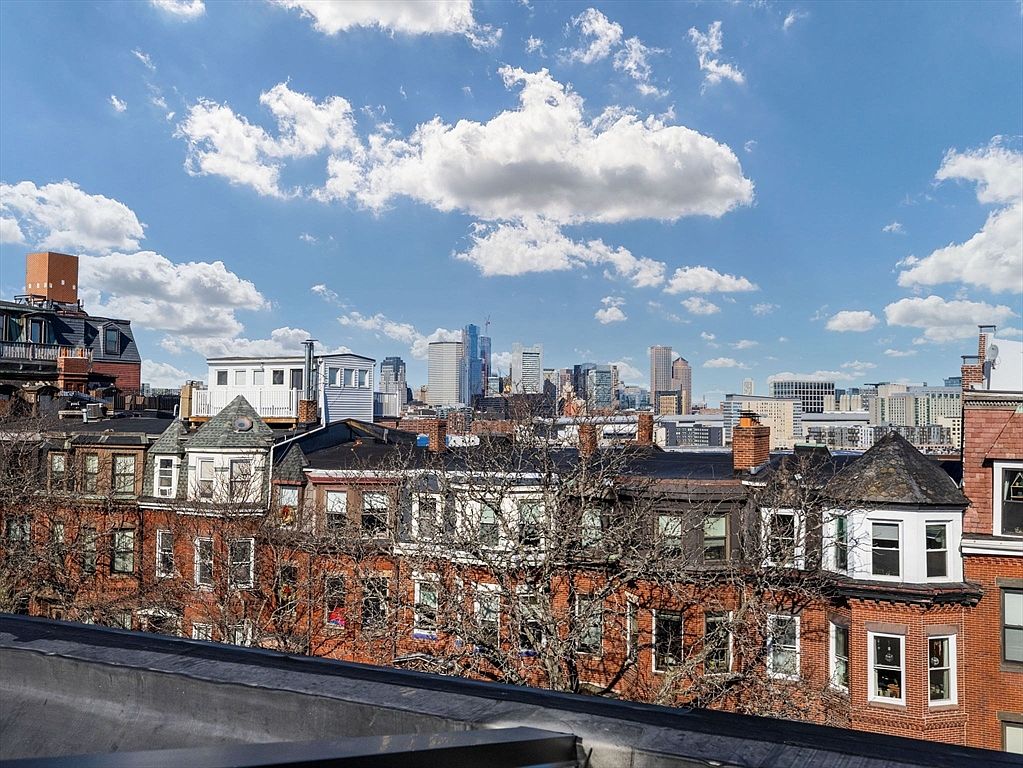 This image showcases an aerial view of a residential neighborhood with brick buildings and a cityscape in the background. The buildings feature a mix of architectural styles, with visible windows, rooftops, and chimneys. The sky is blue with scattered clouds, providing a bright and airy feel to the scene.