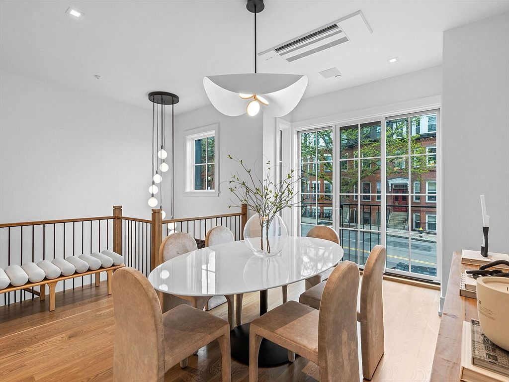 This is a bright and airy dining room featuring a modern aesthetic. The room is furnished with a white oval dining table surrounded by upholstered chairs, and illuminated by two contemporary pendant lights. A large sliding glass door provides ample natural light and a view of the street, while a bench with cushions sits against a railing on the left.