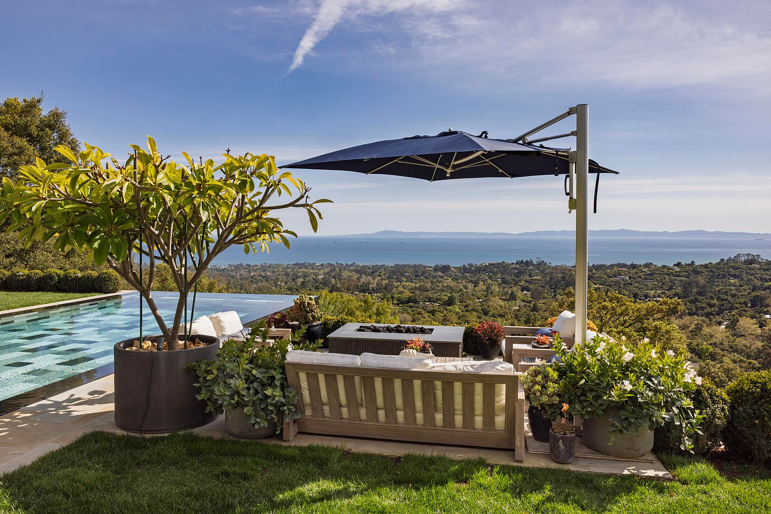This image showcases a luxurious outdoor patio or deck area with stunning panoramic views of the ocean and surrounding landscape. The space is furnished with comfortable seating, including a wooden sofa with cushions, a fire pit, and an umbrella for shade. Lush greenery and potted plants add to the serene and upscale ambiance, making it an ideal spot for relaxation and entertainment.