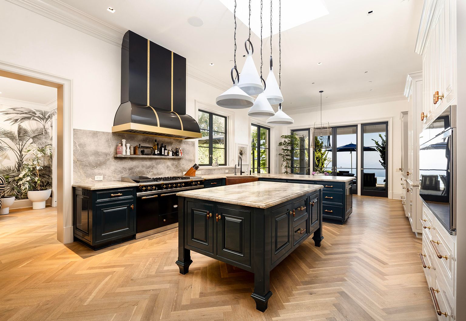 This is a spacious and luxurious kitchen featuring dark blue cabinetry with gold hardware, a large marble-topped island, and a professional-grade range with a custom hood. The herringbone wood flooring adds warmth and elegance, while the pendant lighting and natural light from the windows create a bright and inviting atmosphere. The perspective is a wide shot, showcasing the entire kitchen layout and its high-end finishes.