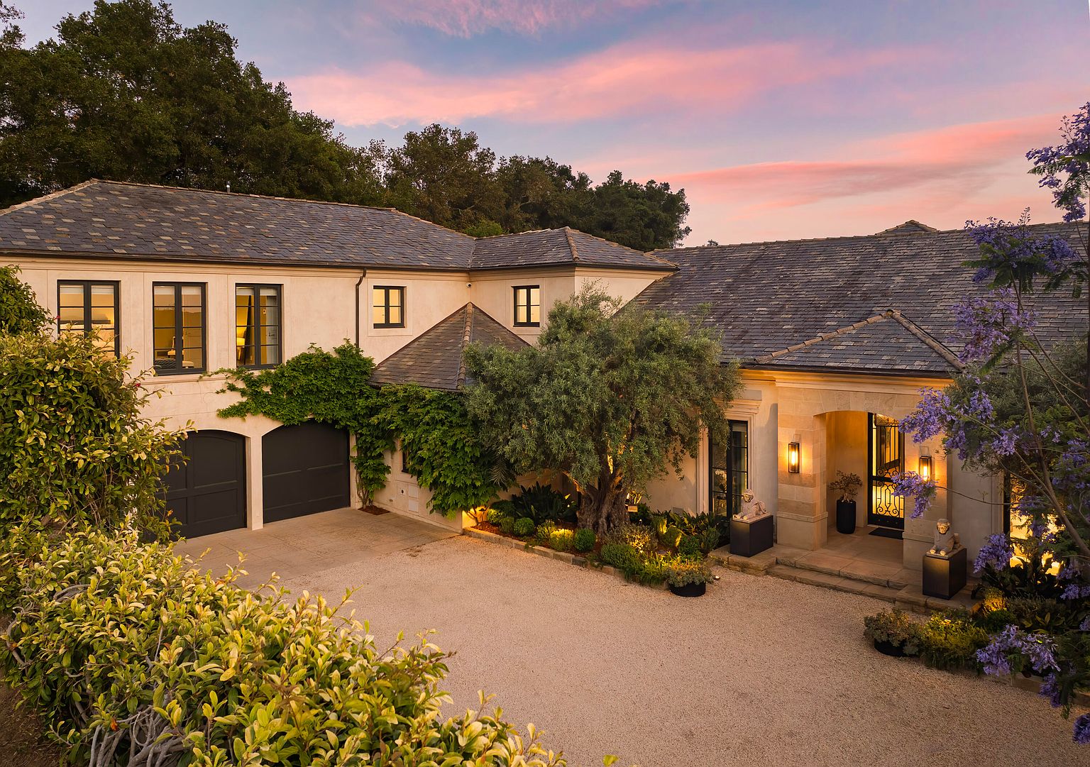 This is a front exterior view of a luxurious home featuring a stone facade, a dark gray roof, and black-framed windows. The property is beautifully landscaped with mature trees, flowering plants, and manicured greenery, creating a welcoming and elegant curb appeal. A gravel driveway leads to the entrance, enhancing the estate's sophisticated charm.