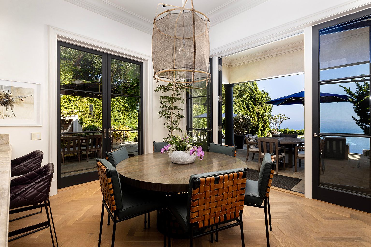 This dining room features a round wooden table surrounded by chairs with woven leather backs and green cushions. A unique, large woven light fixture hangs above the table, and a white bowl with purple orchids serves as a centerpiece. Large glass doors open to an outdoor patio with ocean views, creating a seamless indoor-outdoor living experience.