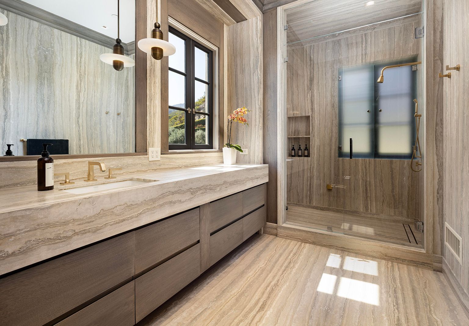 This is a luxurious primary bathroom featuring a long vanity with a marble countertop and dark wood cabinets. A large mirror hangs above the vanity, reflecting pendant lights. A glass-enclosed shower with gold fixtures is visible to the right, and the walls and floors are covered in a matching marble tile, creating a cohesive and elegant design.