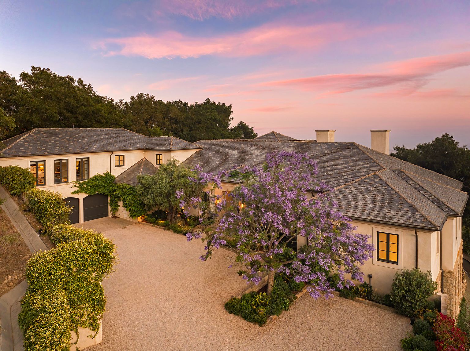 This aerial view showcases a sprawling estate with a meticulously landscaped front yard, featuring a circular driveway and mature trees, including a vibrant purple-blossomed tree. The house itself boasts a neutral color palette, dark-framed windows, and a dark gray roof, exuding a sense of understated elegance and privacy. The surrounding lush greenery adds to the property's secluded and luxurious feel.
