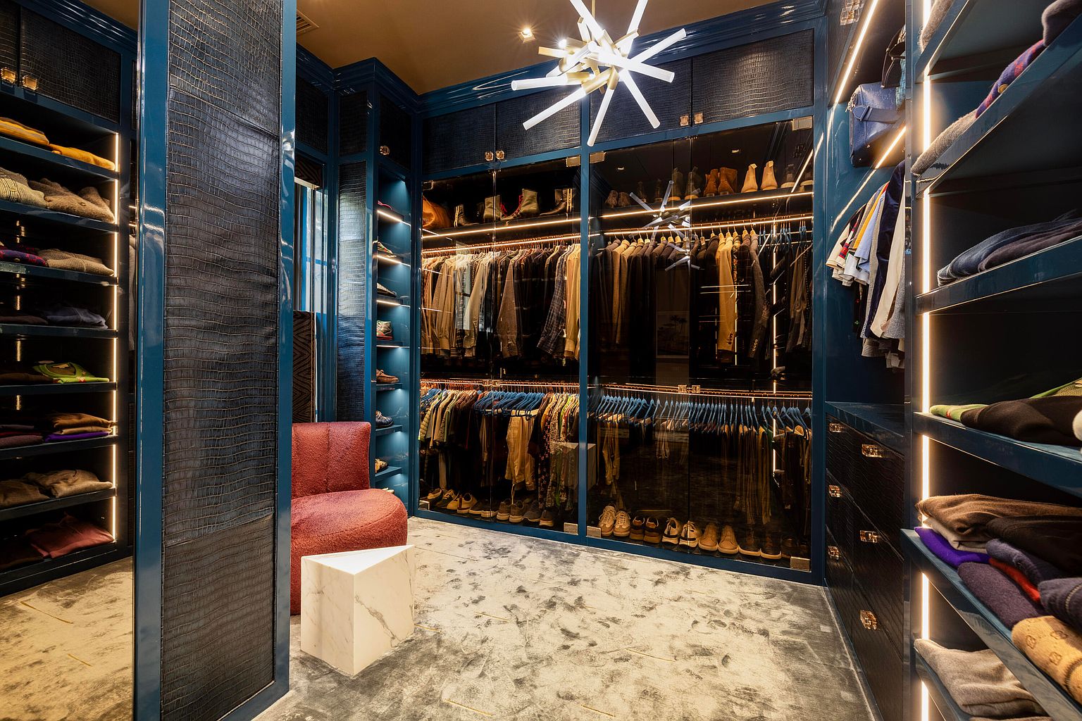 This is a luxurious walk-in closet featuring custom blue cabinetry with glass doors, showcasing an extensive wardrobe. The closet is well-lit with integrated lighting and a modern chandelier, and includes a plush red chair and a marble side table. The overall impression is one of high-end organization and style.