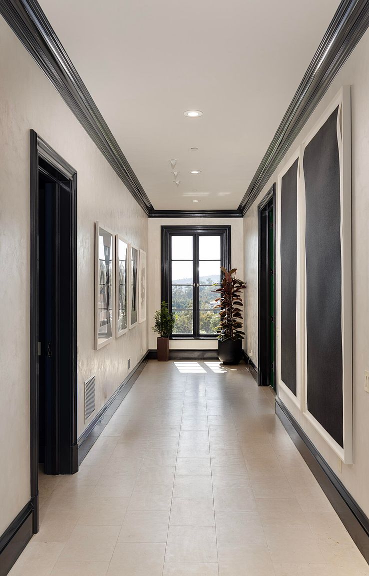 This is a well-lit hallway featuring a neutral color palette with contrasting black trim. The hallway is adorned with artwork and plants, adding a touch of sophistication and life. The flooring appears to be tile, and the overall impression is clean, modern, and elegant.