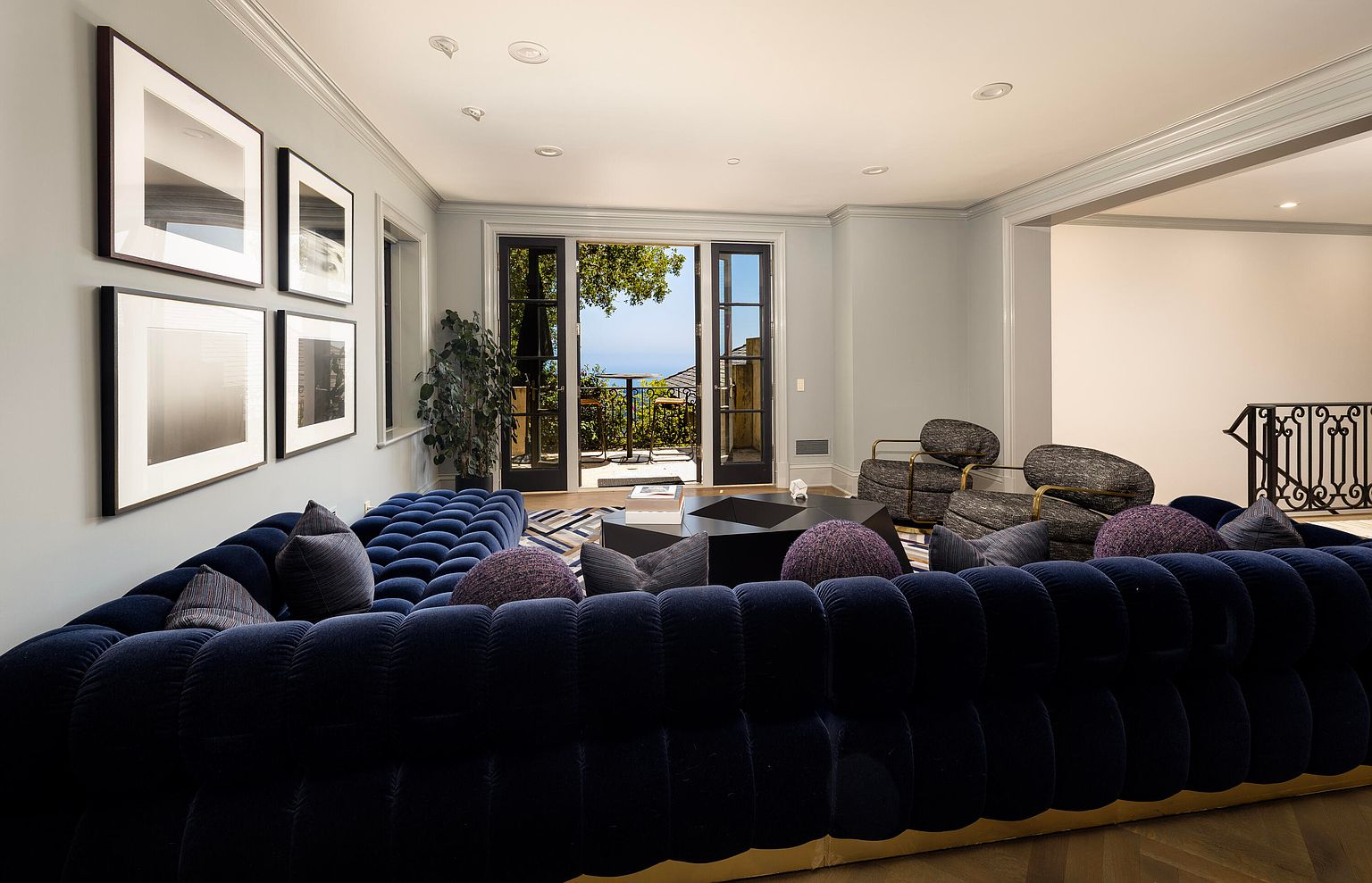 This is an interior shot of a luxurious living room featuring a large, navy blue, tufted sectional sofa with decorative pillows. The room is well-lit with recessed lighting and natural light coming through the open balcony doors, which offer a glimpse of the outdoor scenery. The space also includes modern armchairs, a geometric coffee table, and framed artwork on the walls, creating an elegant and inviting atmosphere.