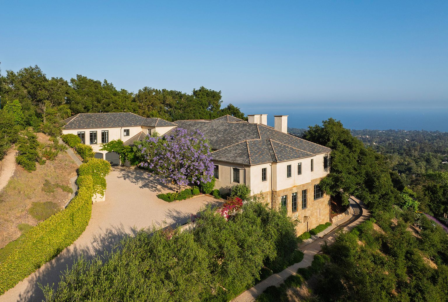 This aerial view showcases a luxurious estate nestled on a hillside, featuring a sprawling residence with a stone and stucco facade, a gray tile roof, and multiple chimneys. The property is surrounded by lush greenery, mature trees, and meticulously landscaped gardens, with a winding driveway leading to the house. In the background, the ocean provides a stunning backdrop, enhancing the property's appeal and exclusivity.