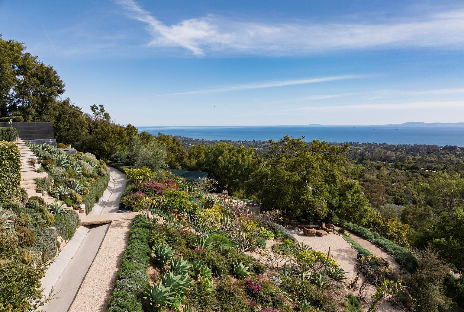 This image showcases a beautifully landscaped yard and garden with a stunning ocean view. The garden features a variety of plants, including succulents and flowering shrubs, arranged along pathways and terraces. A concrete aqueduct runs through the garden, adding a unique architectural element. The overall impression is one of tranquility and natural beauty, perfect for a luxury real estate listing.