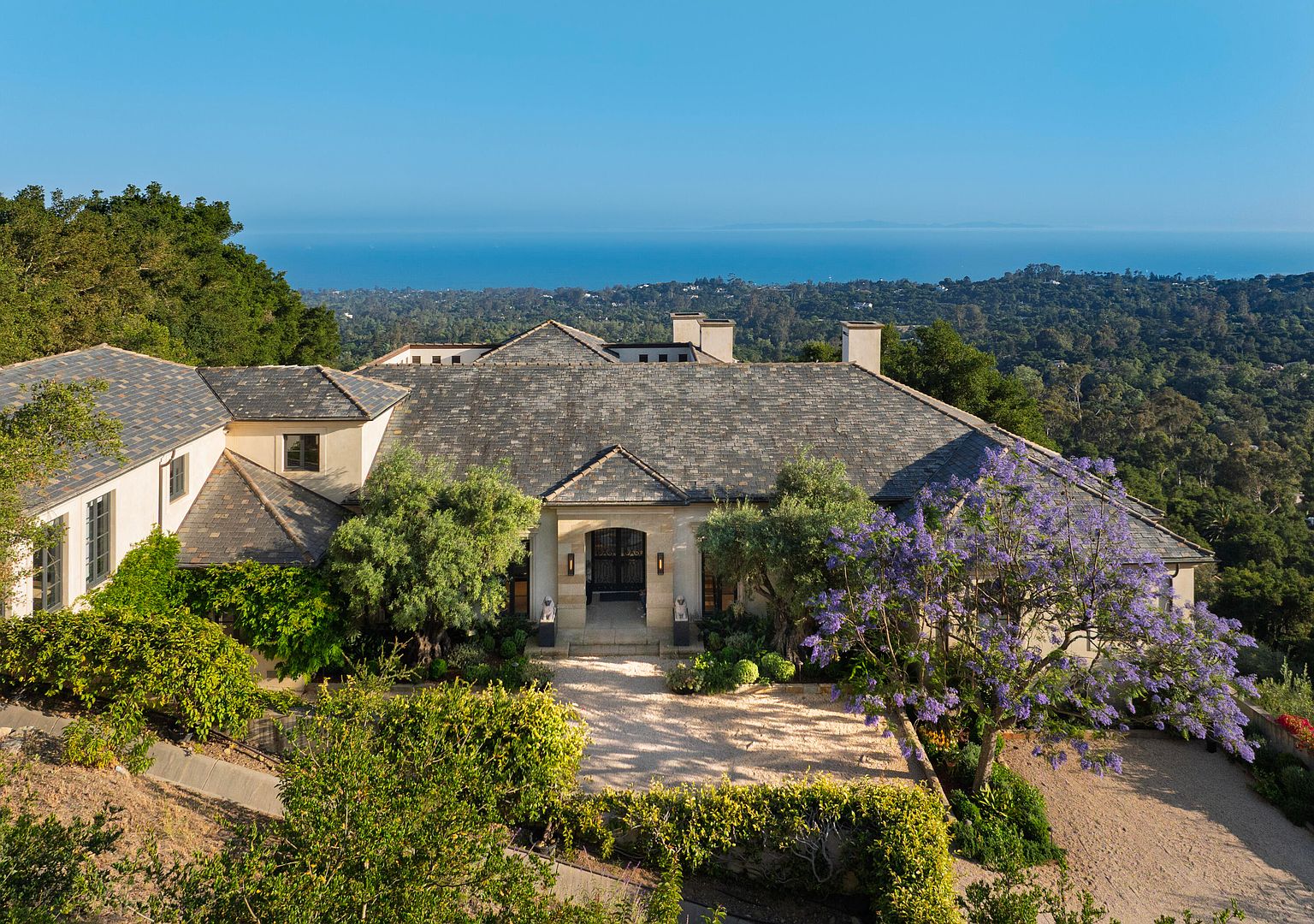 This is a front exterior view of a luxurious estate. The house features a stone roof, a grand entrance with stone pillars, and meticulously landscaped grounds with mature trees and flowering plants. The property offers stunning views of the ocean and surrounding landscape, creating a sense of privacy and exclusivity.