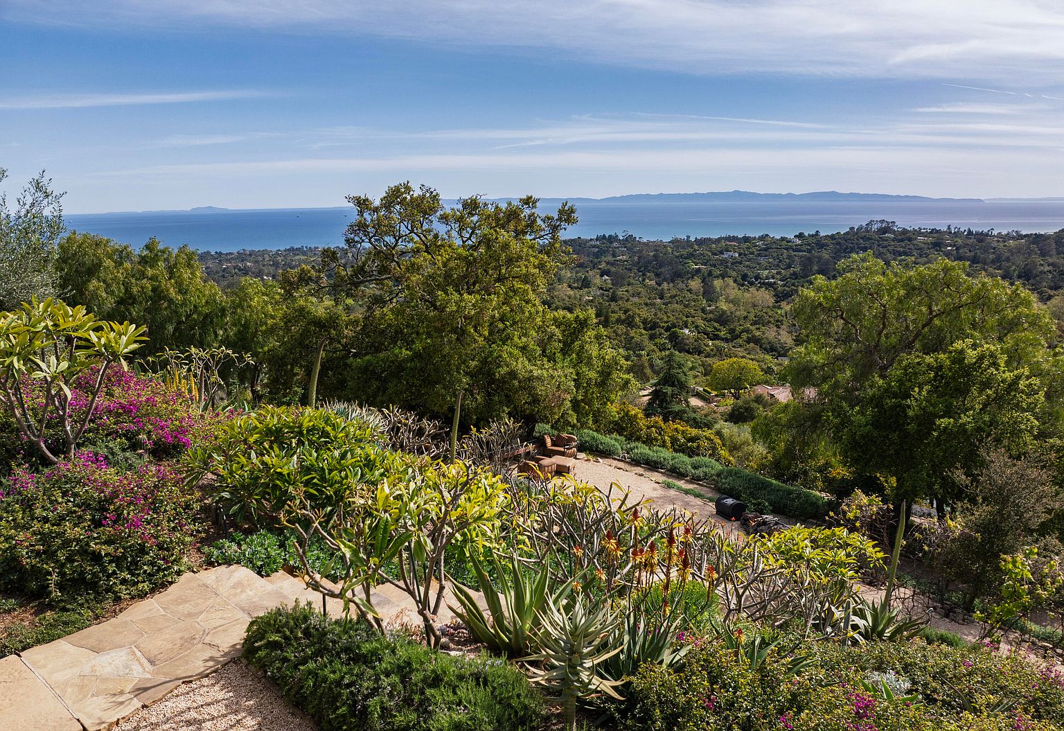This image showcases a beautifully landscaped yard and garden with a stunning ocean view. The foreground features lush greenery, colorful flowers, and stone pathways, leading to a panoramic vista of the ocean and distant islands. The scene evokes a sense of tranquility and natural beauty, highlighting the property's outdoor living potential.