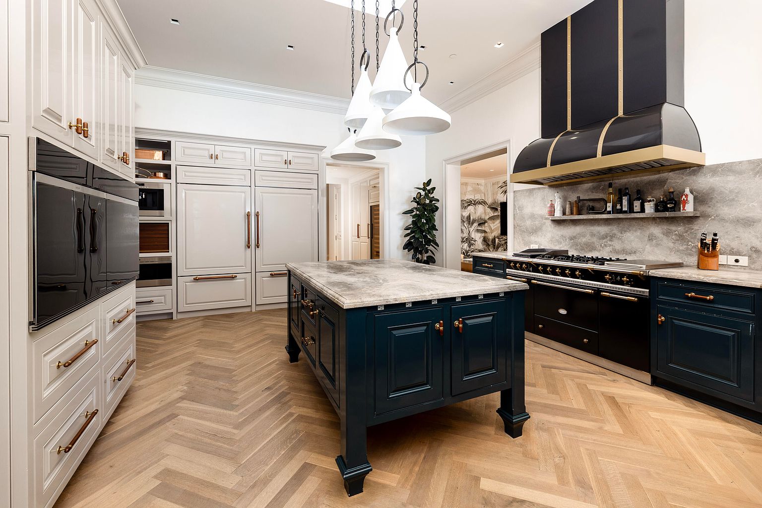This is a well-lit, high-end kitchen featuring custom cabinetry in white and dark blue, complemented by copper hardware. A large center island with a stone countertop anchors the space, while professional-grade appliances and a statement range hood add to the luxurious feel. The herringbone wood flooring and pendant lighting contribute to the kitchen's elegant and inviting atmosphere.