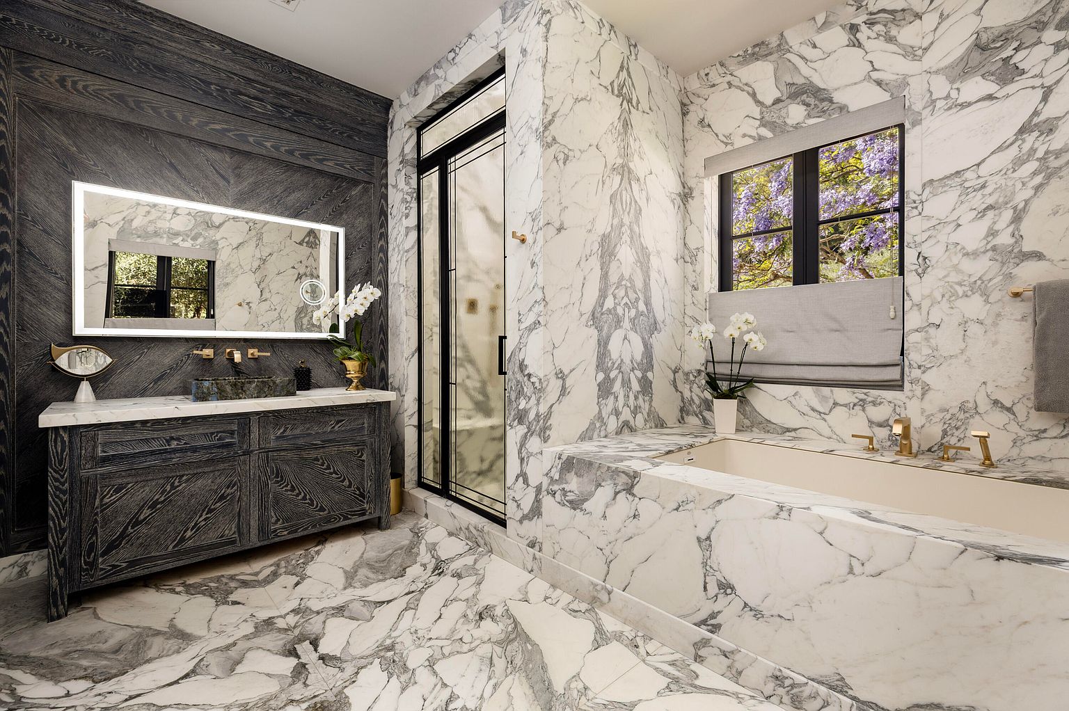 This is a luxurious primary bathroom featuring marble walls and flooring, creating a spa-like atmosphere. A modern vanity with a unique vessel sink and illuminated mirror is complemented by a freestanding bathtub surrounded by marble. A glass-enclosed shower adds to the upscale feel of the space.