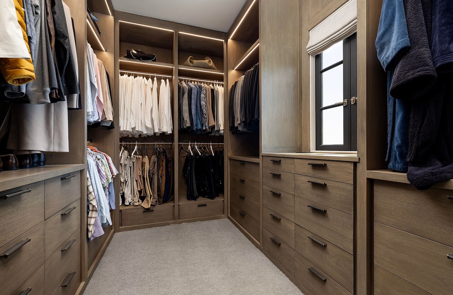 This is a well-organized walk-in closet featuring custom wood cabinetry with built-in drawers and shelving. Clothes are neatly hung and folded, and the closet is illuminated by recessed lighting. A window provides natural light, enhancing the luxurious feel of the space.