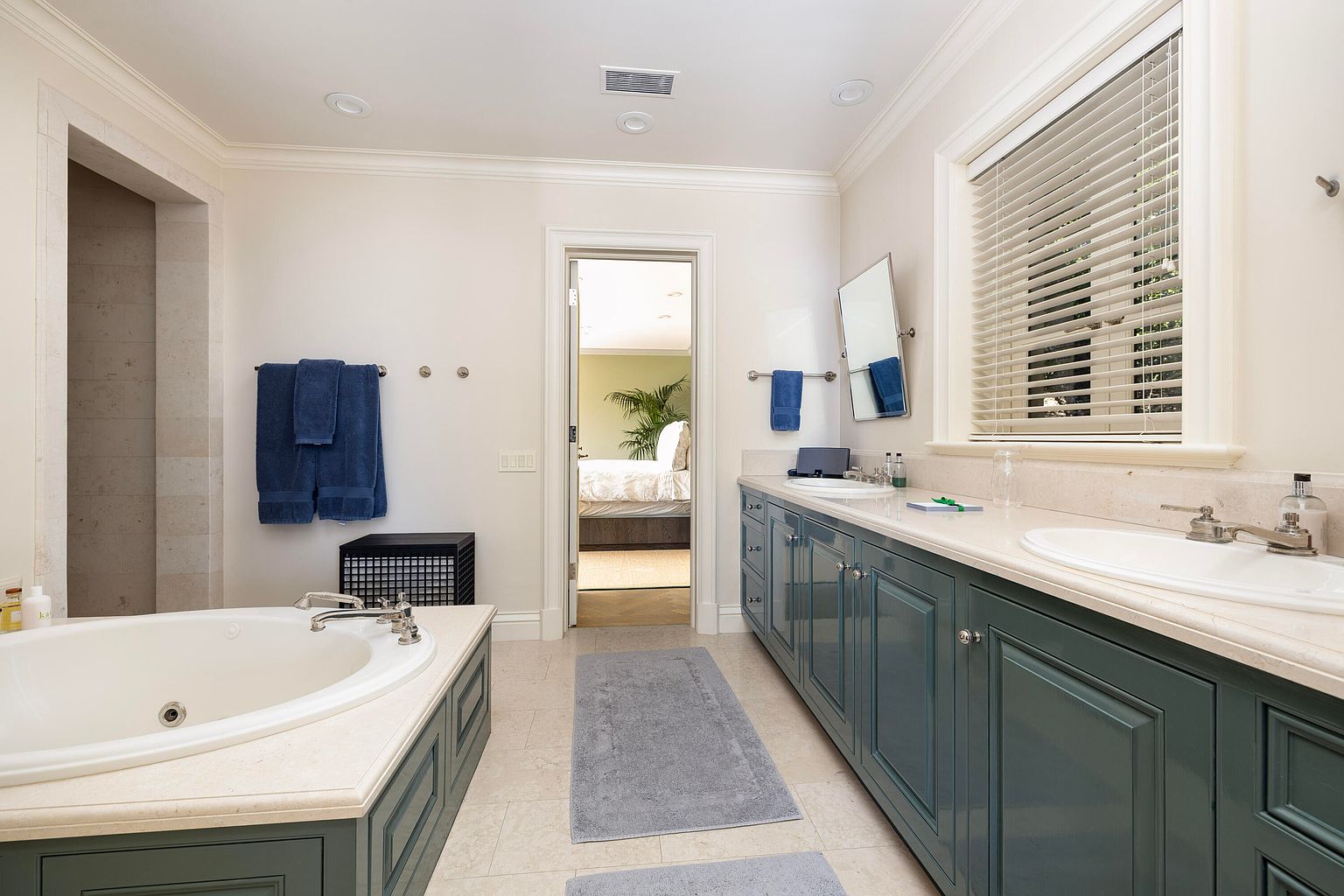 This is a well-lit primary bathroom featuring a large soaking tub with a blue-green paneled base, a double vanity with matching blue-green cabinetry and light countertops, and a separate shower area. The room has neutral-toned walls and tile flooring, with blue towels adding a pop of color. The bathroom opens into a bedroom, creating a sense of spaciousness and luxury.