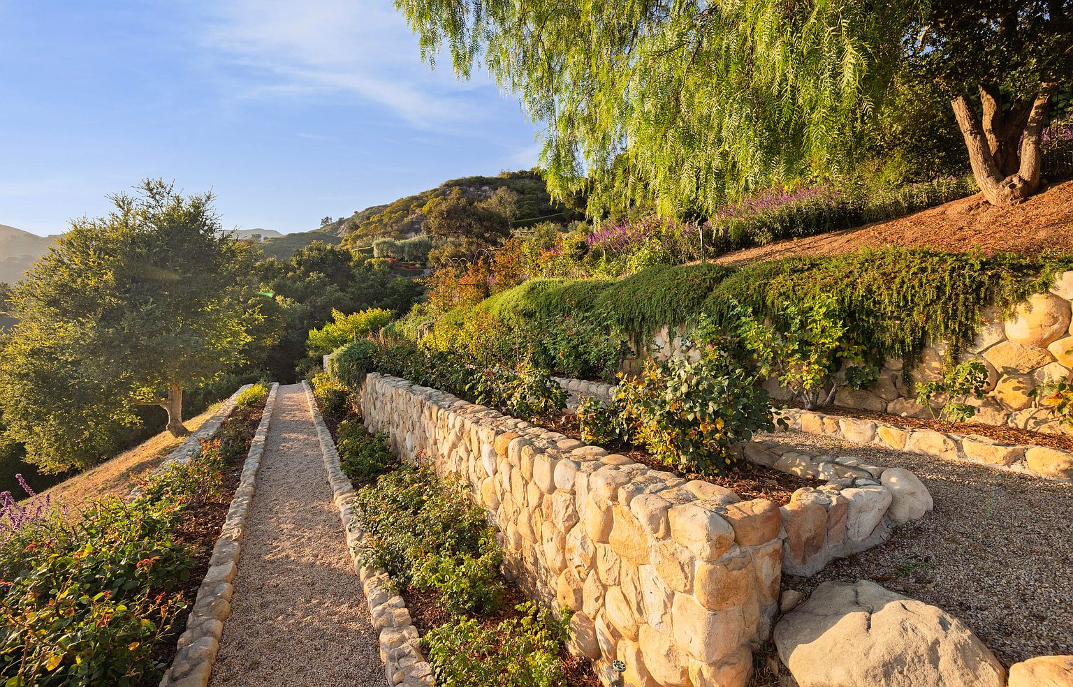 This image showcases a beautifully landscaped yard or garden, featuring stone retaining walls, gravel pathways, and lush greenery. The tiered design adds visual interest, while the variety of plants and flowers creates a serene and inviting atmosphere. The overall impression is one of tranquility and meticulous care, highlighting the property's outdoor appeal.