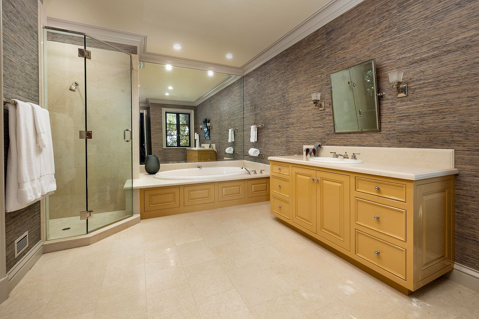 This is a well-lit primary bathroom featuring a glass-enclosed shower, a large soaking tub with wood paneling, and a vanity with a light-colored countertop and cabinetry. The walls are covered in a textured wallpaper, and a large mirror spans the wall above the tub. The flooring is a light-colored tile, creating a clean and luxurious atmosphere.