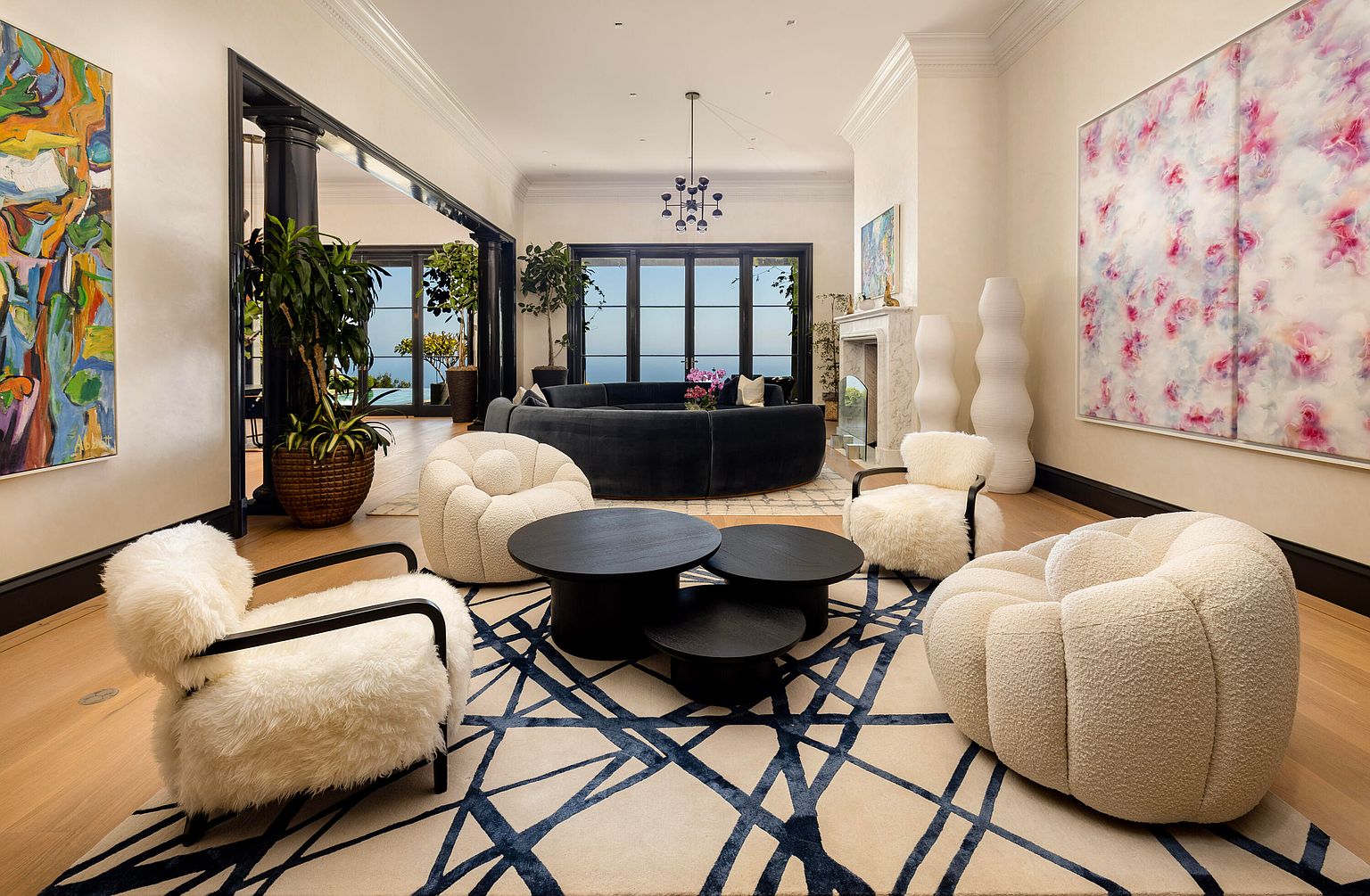 This is a luxurious living room featuring a curved sofa, plush armchairs, and a unique geometric rug. The room is bright and airy, with large windows offering a view of the ocean. The space is decorated with modern art and stylish accessories, creating a sophisticated and inviting atmosphere.