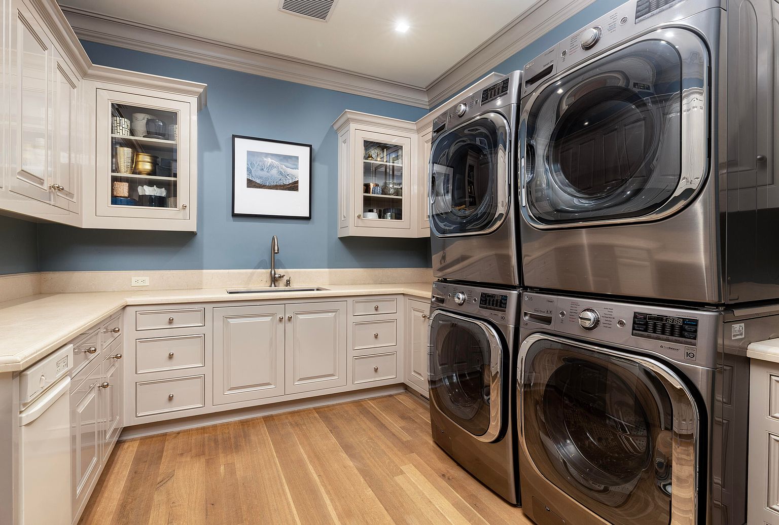 This is a well-appointed laundry room featuring stacked, stainless steel washer and dryer units. The room includes custom cabinetry in a neutral tone, complemented by a light countertop and a sink. The walls are painted a calming blue, and the hardwood floors add warmth to the space, creating a functional yet stylish area.