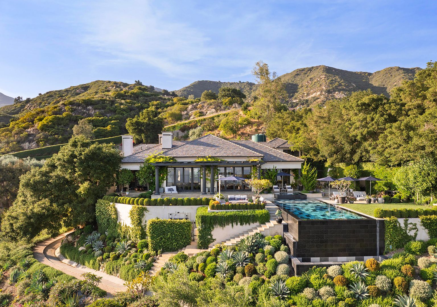 This aerial view showcases a luxurious estate nestled in a lush, mountainous landscape. The property features a modern house with a dark roof, a stunning infinity pool with a waterfall feature, and meticulously landscaped gardens with terraced levels. A winding driveway leads to the house, emphasizing the privacy and exclusivity of the location.