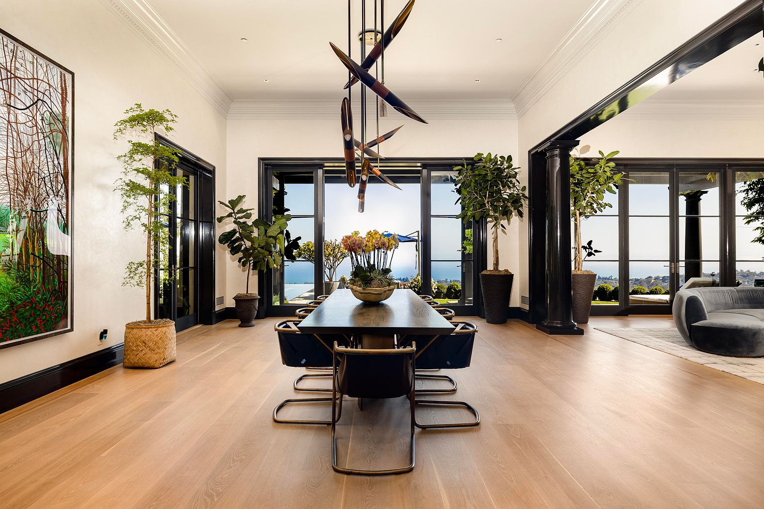 This is a spacious dining room featuring a long, dark wood table with modern chairs, illuminated by a unique, sculptural chandelier. Large windows and doors offer views of the ocean and landscape, enhancing the room's bright and airy atmosphere. The room is decorated with potted plants and artwork, adding a touch of elegance and sophistication.