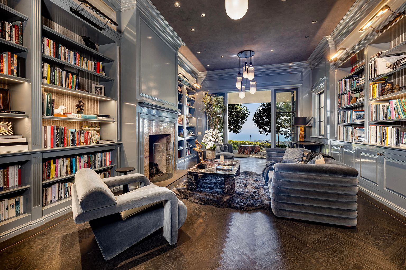This is a luxurious living room featuring custom built-in bookshelves filled with books and decorative items. The room is furnished with plush velvet seating, a unique coffee table, and a statement lighting fixture. Large windows offer a view of the outdoors, creating a sophisticated and inviting atmosphere.