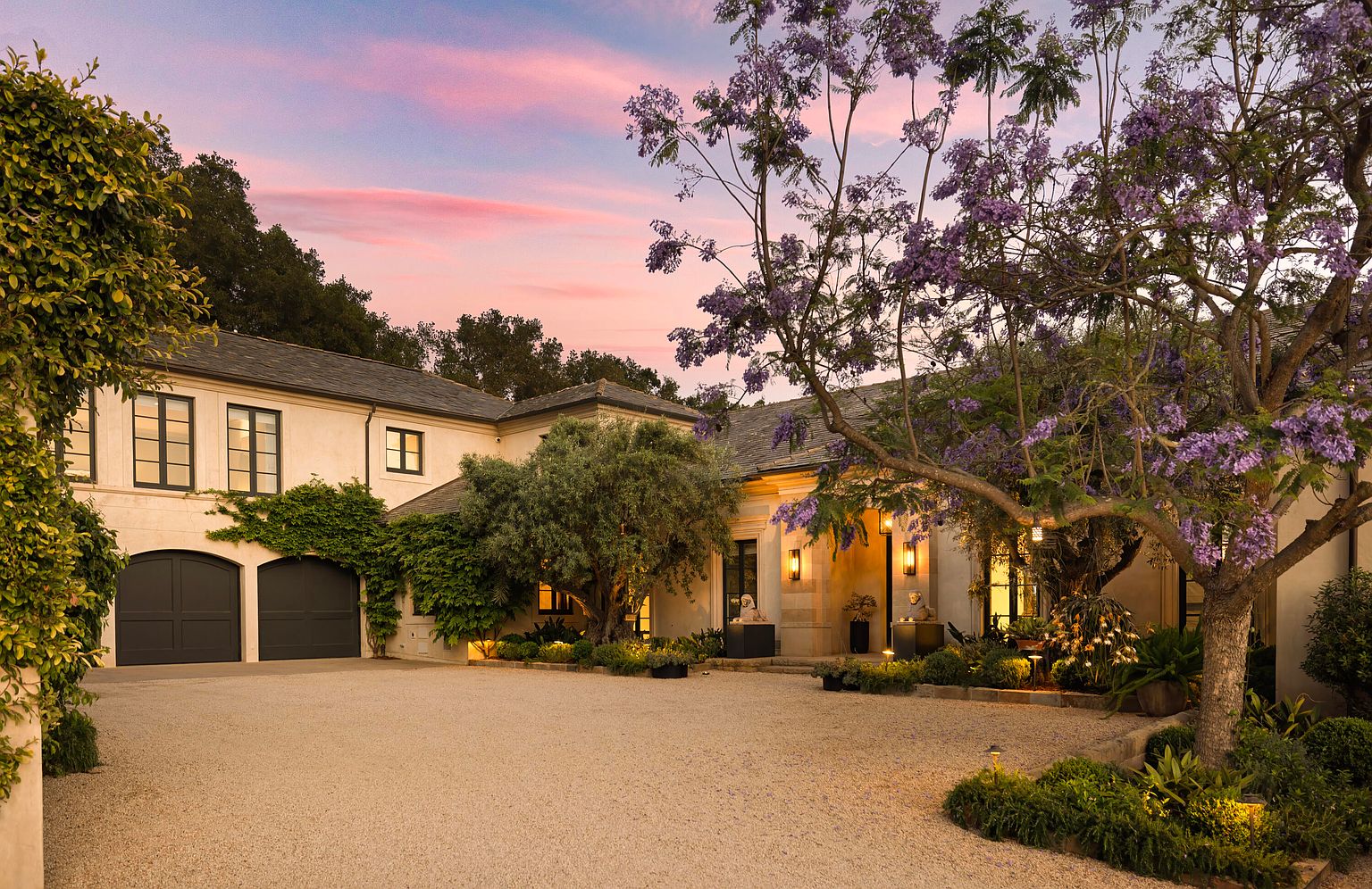This is a front exterior view of a luxurious home at dusk. The house features a light-colored facade, dark-framed windows, and a dark gray roof. A gravel driveway leads up to the house, which is surrounded by lush landscaping, including mature trees with purple flowers and climbing vines. Warm lighting emanates from the entryway, creating an inviting atmosphere.