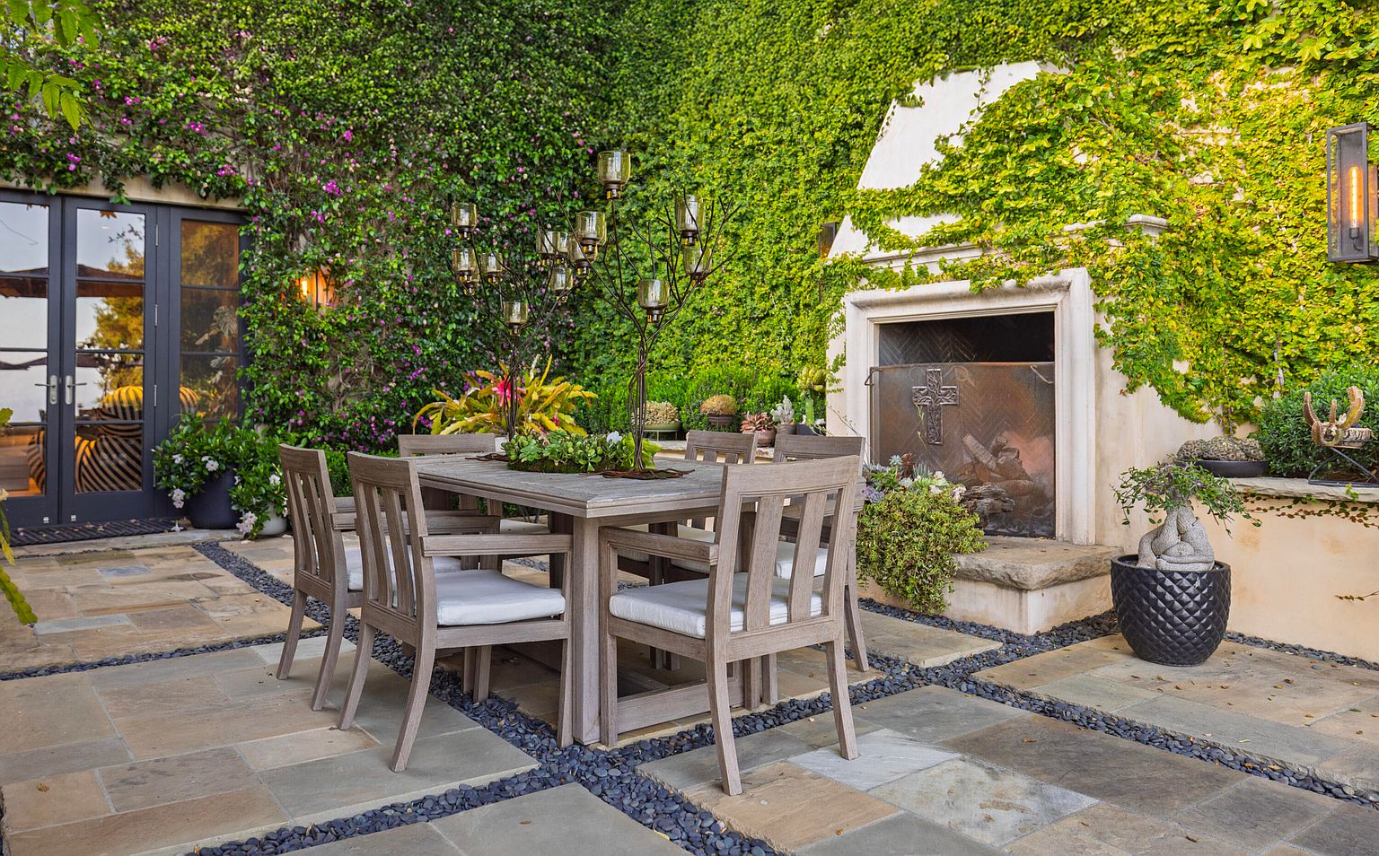 This image showcases an inviting outdoor patio area, featuring a wooden dining set with cushioned chairs arranged on a stone-tiled surface accented with dark gravel. Lush greenery, including climbing vines covering the walls and potted plants, creates a serene and private atmosphere, while a unique fireplace adds a focal point and architectural interest. The perspective is at eye level, creating a sense of immersion in the outdoor space.