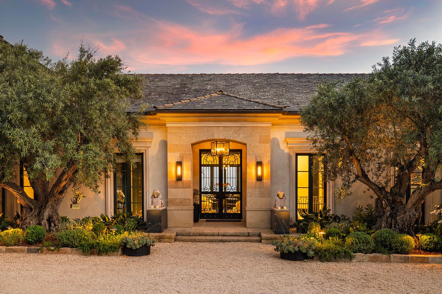 This is a stunning entryway to a luxurious home, featuring a grand black iron gate framed by stone pillars and illuminated by elegant sconces. Mature olive trees flank the entrance, adding to the property's curb appeal. The gravel driveway and manicured landscaping create a welcoming and sophisticated ambiance, perfect for a high-end real estate listing.
