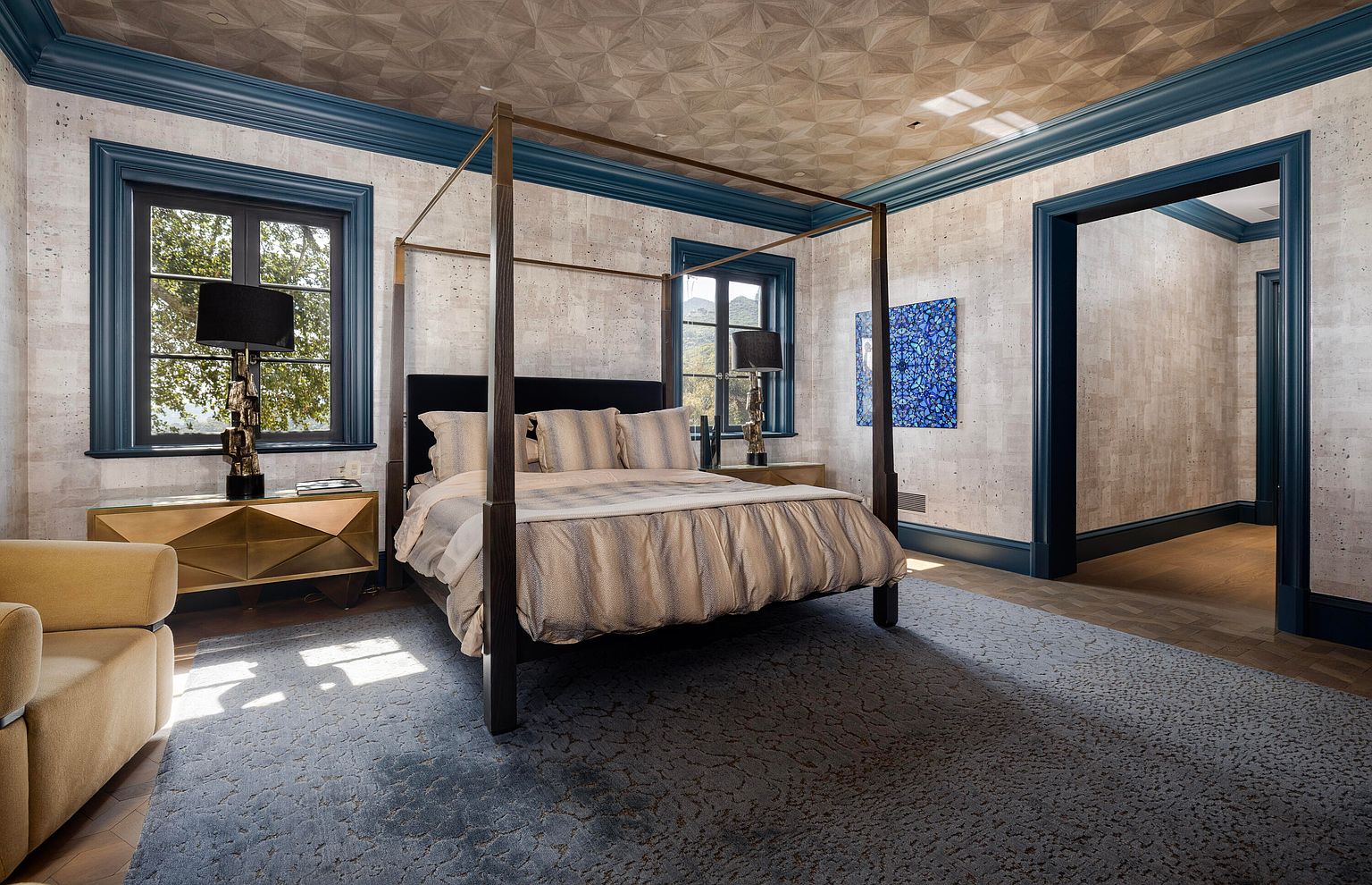 This is a luxurious primary bedroom featuring a four-poster bed with elegant bedding, complemented by matching nightstands and lamps. The room is decorated with textured wallpaper, a patterned ceiling, and striking blue trim, creating a sophisticated and inviting atmosphere. A blue abstract painting adds a pop of color, and the room is bathed in natural light from the windows.