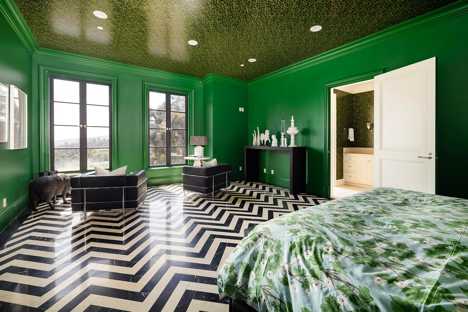This is a primary bedroom featuring bold green walls and a patterned ceiling, creating a unique and luxurious atmosphere. The room includes large windows, black and white chevron flooring, and a glimpse into an adjacent bathroom. The bed is covered with a floral patterned duvet, adding a touch of softness to the striking design.