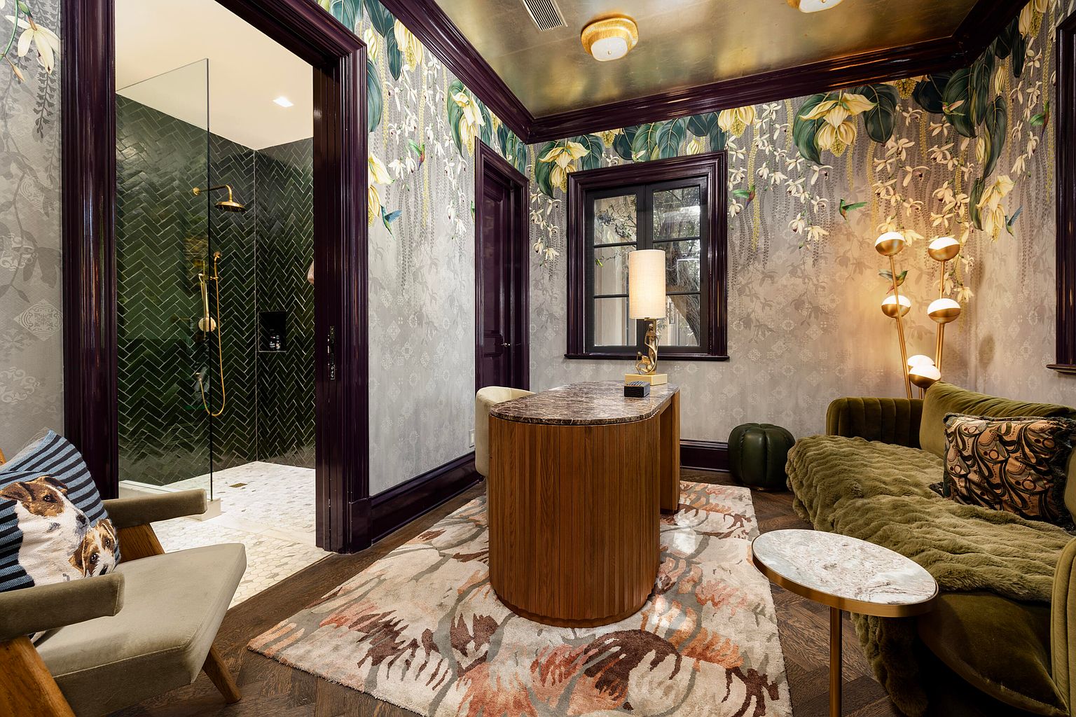 This interior shot showcases a luxurious office or study with a unique design. The room features floral wallpaper, a custom desk, and a comfortable green velvet sofa. A glimpse into an adjacent bathroom with green herringbone tiles adds an unexpected touch, creating a sophisticated and inviting workspace.