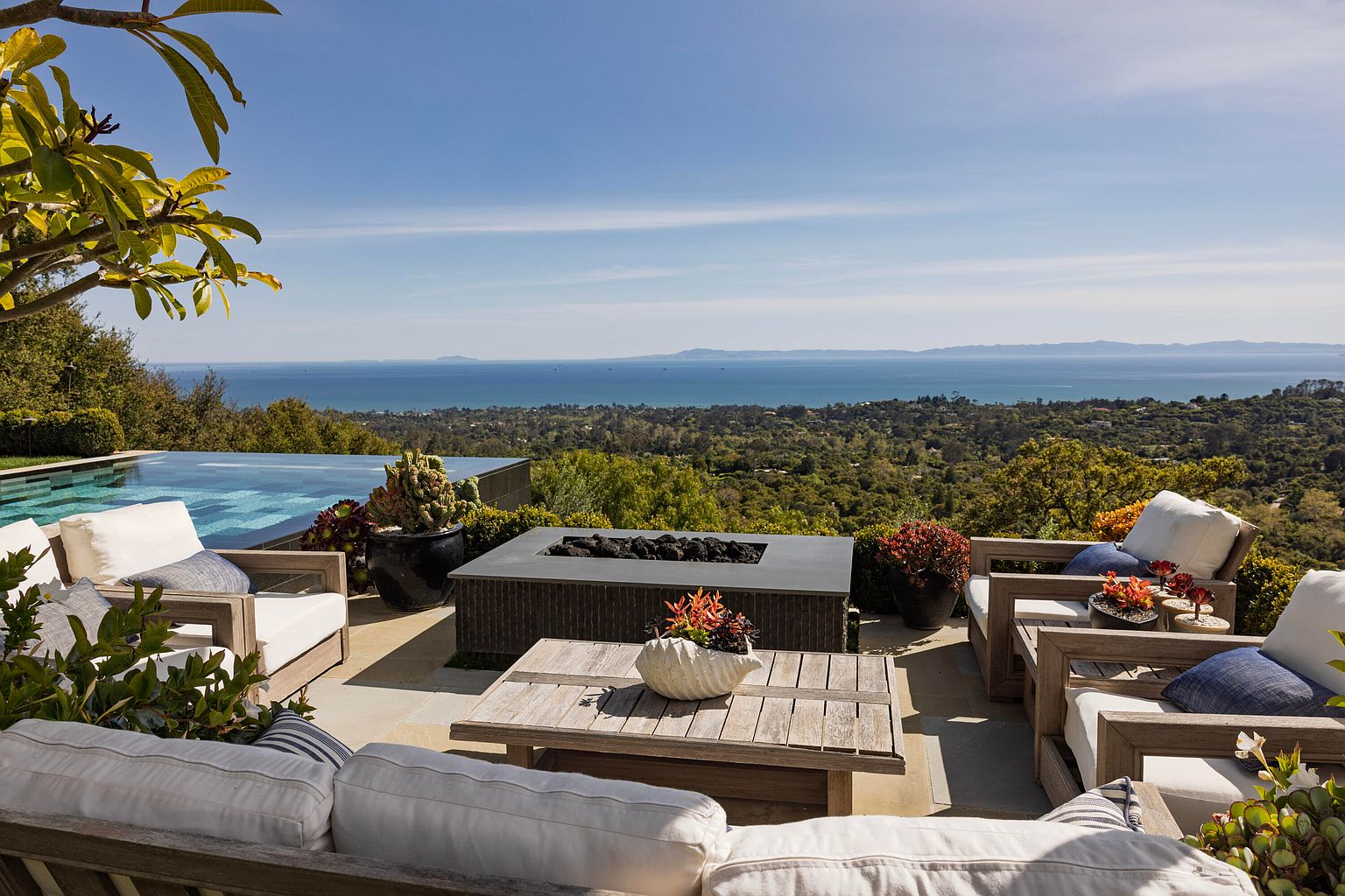 This image showcases a luxurious outdoor patio area with stunning ocean views. The space is furnished with comfortable seating arrangements around a modern fire pit and a rustic wooden coffee table. An infinity pool is visible on the left, blending seamlessly with the horizon, creating a serene and upscale ambiance perfect for relaxation and entertainment.