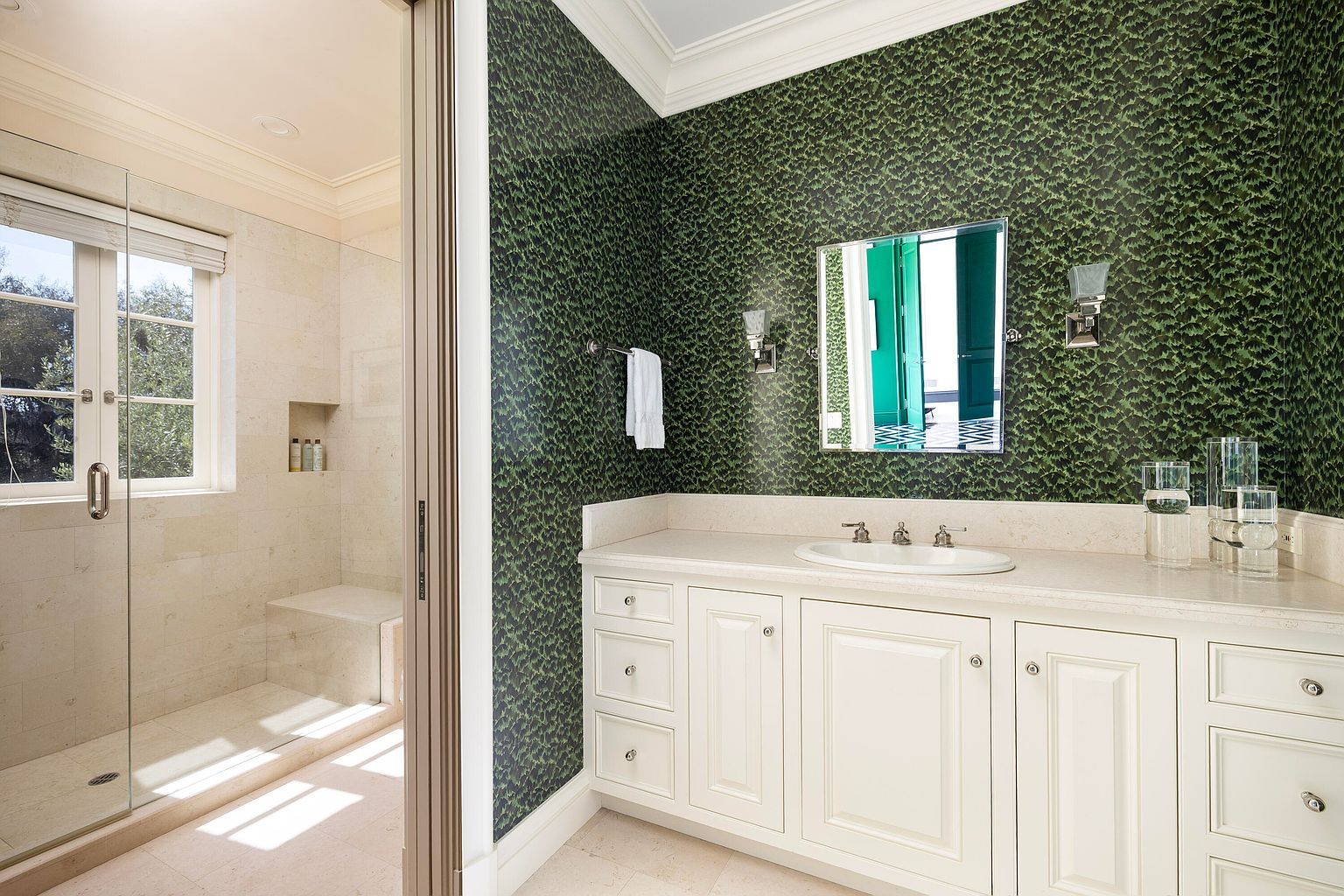 This is a well-appointed bathroom featuring a walk-in shower with glass doors and a built-in bench. The vanity has a white cabinet with a light countertop, complemented by green patterned wallpaper. The overall design is elegant and clean, creating a relaxing atmosphere.