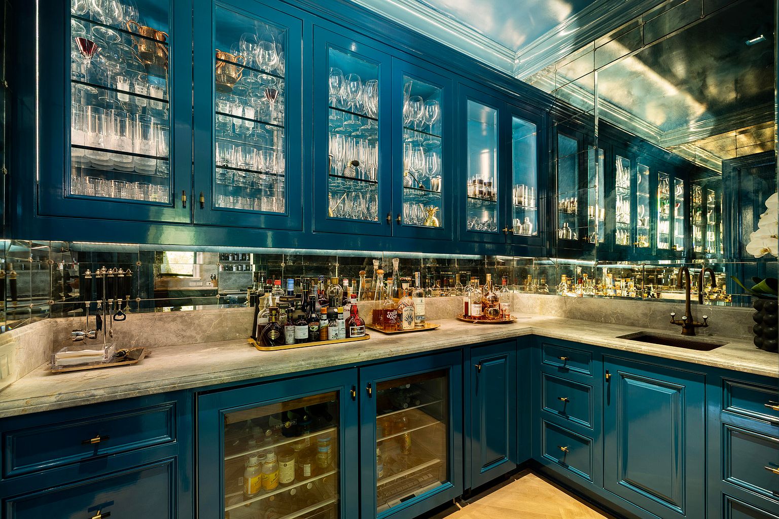This is an interior shot of a sophisticated wine cellar featuring custom blue cabinetry with glass doors displaying various glasses and bottles. The countertop is a light-colored stone, and the backsplash is mirrored, adding depth and elegance to the space. The overall impression is luxurious and well-appointed, perfect for showcasing a curated wine collection.